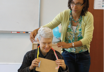 Michele is reaching for a prototype from a blind research participant. The artifact is a clip board hanging from the participant's neck using pipe cleaners taped to the board.