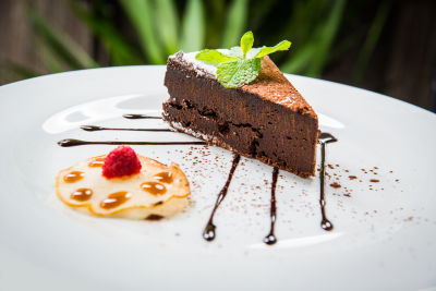 A slice of flourless chocolate cake plated slightly off center, garnished with cocoa powder, chocolate drizzle, and a sprig of mint.