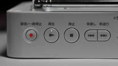 Close-up of a player showing physical playback control buttons: record (red dot), play (right-pointing triangle), stop (square), rewind (double left arrows), and fast forward (double right arrows). Japanese labels appear above each button. (Note: The pause button (two vertical bars) is not shown in the picture.)