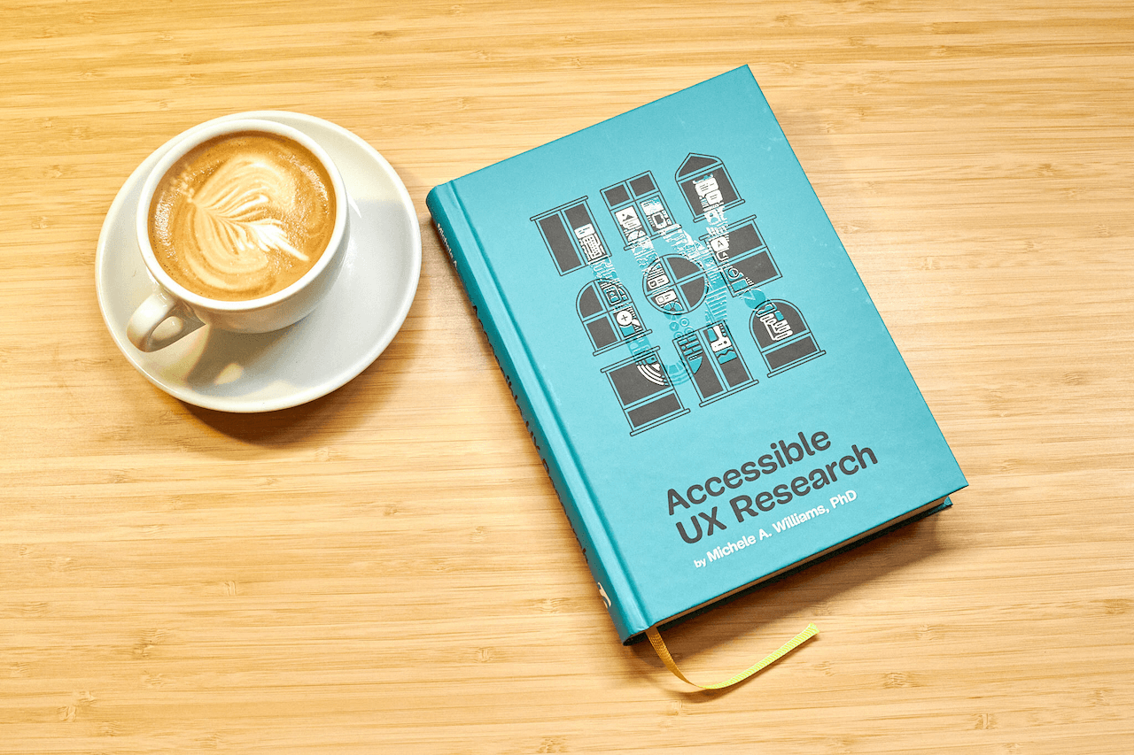 Hardcover edition of Michele Williams&rsquo; new book &lsquo;Accessible UX Research&rsquo; on a wooden table, with a cup of coffee next to it. The book has a teal cover that shows a three times three grid of windows in different architectural styles. On top of the windows, there&rsquo;s a light varnish that spells out the word &lsquo;UX&rsquo; with research-related icons such as speech bubbles, a looking glass, a keyboard, and UI components.